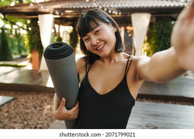 Asian woman taking selfie photo while standing with yoga mat outdoors