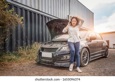 Asiatische Frau, die mit Smartphone in der Nähe von defektem Auto spricht, mit geöffneter Motorhaube und Stress zeigen.
