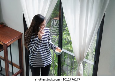 Asian Woman Talking On Mobile Phone And Standing At Cafe Restaurant Near Window See Through Garden,Digital Age Lifestyle,Working Outside Office Concept.