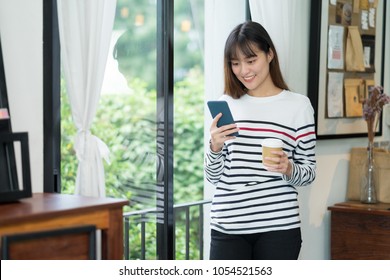 Asian Woman Talking On Mobile Phone Video Call And Standing At Cafe Restaurant Near Window See Through Garden,Digital Age Lifestyle,Working Outside Office Concept.