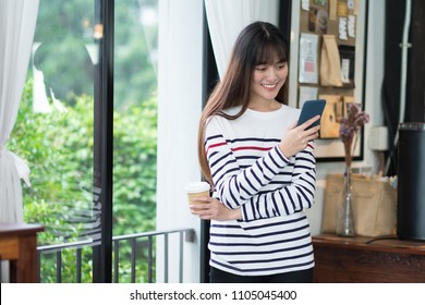 Asian Woman Talking On Mobile Phone Video Call And Standing At Cafe Restaurant Near Window See Through Garden,Digital Age Lifestyle,Working Outside Office Concept.