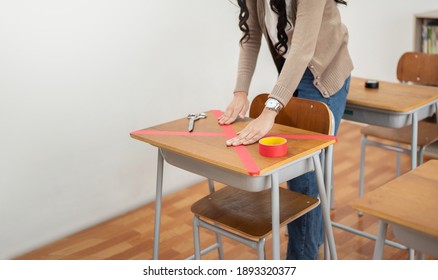 Asian Woman Teacher Applying Marking Red Duct Tape In Crossed Symbol On Desk In Classroom At School During Covid-19 Pandemic.