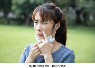 Asian Woman Tiring Of Mask And Removing Face Mask In Outdoor Park Due To Acne Or Pimple On Her Face, Concept Of Lockdown Fatigue, Negative Effect Of Day Long Face Mask Wearing