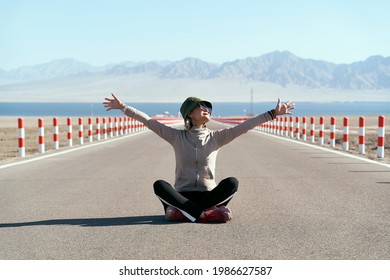 asian woman tourist sitting in the middle of an empty open road with lake and rolling mountains in background, leg crossed arms outstretched. - Shutterstock ID 1986627587