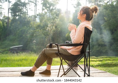 Asian woman tourist sitting on outdoor chair at mountain resort balcony and drinking a cup of hot coffee in autumn morning. Pretty girl relax and enjoy outdoor lifestyle and holiday travel vacation.