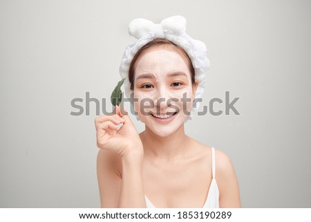 Asian woman in towel applying white facial clay mask and holding a leaf.