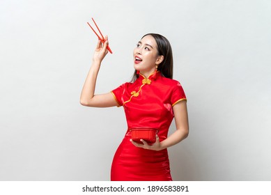 Asian woman in traditional red Chinese qipao dress holding plastic food box and chopsticks looking upward in studio isolated light gray background