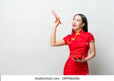 Asian woman in traditional red Chinese qipao dress holding plastic food box and chopsticks looking upward to empty space in studio isolated light gray background