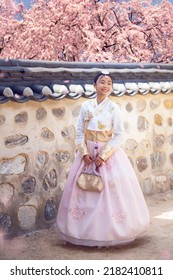 Asian Woman Traveler In Traditional Korean Dress Or Hanbok Dress Walking In Old Palace With Sakura Flower Background, Seoul City, South Korea