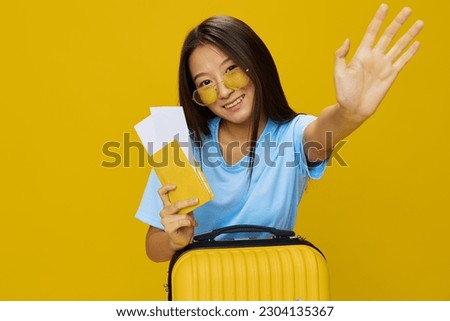 Asian woman traveling with yellow suitcase and tickets with passport in hand, tourist traveling by plane and train with luggage on yellow background in blue T-shirt and jeans