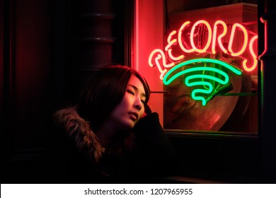 Asian Woman In Trendy Apparel And Sweater Coat Looking Up Enjoying Nightlife In City. Outdoors On Street With Neon City Illumination. Bar And Music Concept