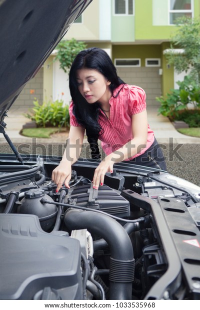Asian woman trying to fix broken down car in front\
of her house