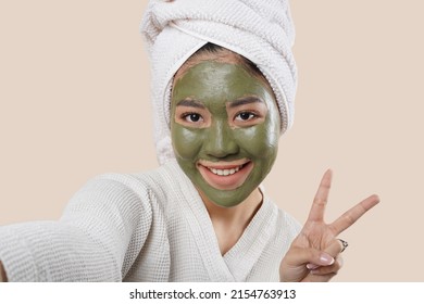 An Asian Woman Using Bathrobe, Towel In Her Hair, And Face Mask, Taking Selfie And Pose To The Camera. Isolated On Cream Background.