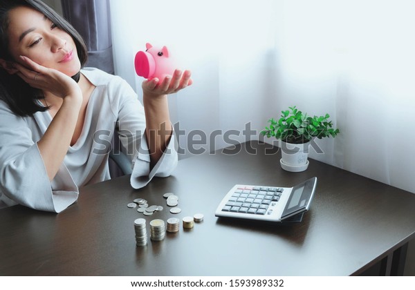 Asian woman using hand on chin or cheek. Smile with\
happiness look at the piggy bank. On the table  have stack of coins\
already counted and a calculator. Successful savings will make\
dreams come true.