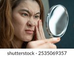 Asian woman using mirror to see acne and wrinkle problem on her face. Allergies, irritants, genetic makeup, etc can cause skin conditions.