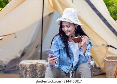 Asian Woman Is Using Mobile Phone While Drinking Coffee At Her Tent While Camping Outdoor During Summer Time In National Park For Adventure And Active Travel