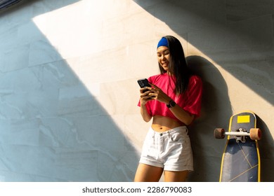 Asian woman using mobile phone with internet during skating on longboard skate on summer vacation. Stylish youth people enjoy urban outdoor lifestyle extreme sports and portable device in the city. - Powered by Shutterstock