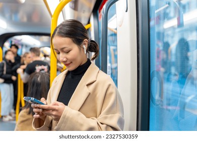 Asian woman using mobile phone and listening to the music on earphones during travel on train in the city. Attractive girl enjoy urban lifestyle with using digital gadget device and online network.
