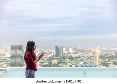 Asian Woman Using New Mobile Application Standing At Infinity Swimming Pool On Rooftop With Skyscrapers In The Background. Woman Looking At Free Space For Your Advertising Content