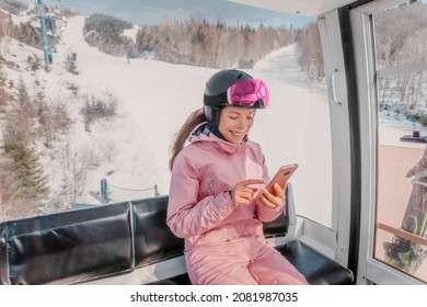 Asian woman using phone on ski holidays - Woman skier using phone app in gondola ski lift. Girl smiling looking at mobile smartphone wearing ski clothing, helmet and goggles. Winter vacation concept