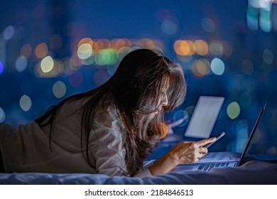 Asian woman using smartphone in her hands, working on laptop, lying on bed near window at home or hotel in the dark at night, warm light from laptop. Blurred background City in night, traffic on road. - Powered by Shutterstock