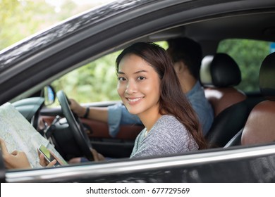 Asian  Woman Using Smartphone And Map And Men Driving Car On Road Trip And Happy Young Couple With A Map In The Car. 