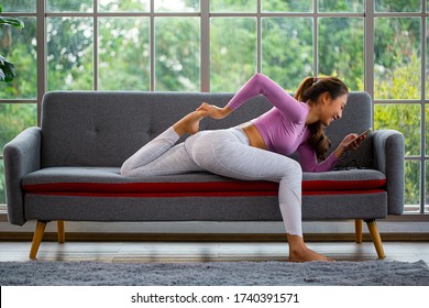 Asian Woman Using Smartphone For Reading New And Check Status And Training Yoga On The Sofa.Thai People Smiling And Relaxing At Home.