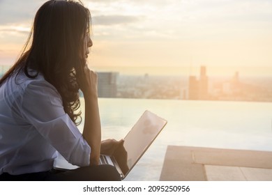 Asian Woman Using Tablet Look In Distance Planning Or Visualizing Future Success, Dreaming Or Thinking, Sitting On Swimming Pool At Sunset. Business Vision Concept.