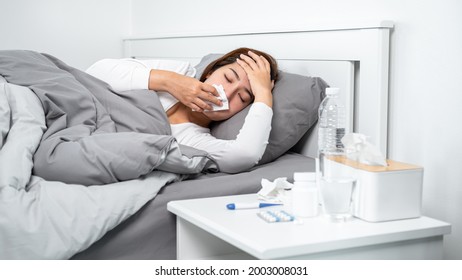 Asian woman using tissue to cough, sneezing and touching forehead while feeling sick and lying in blanket on the bed after using thermometer to checking body temperature and take medicine in bedroom - Shutterstock ID 2003008031