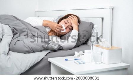 Asian woman using tissue to cough, sneezing and touching forehead while feeling sick and lying in blanket on the bed after using thermometer to checking body temperature and take medicine in bedroom