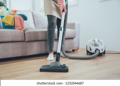 Asian Woman Using Vacuum Cleaner In The Living Room. Housekeeper Wearing Pink Protective Rubber Gloves Cleaning The Wooden Floor In The House. Full Of Clean Supplies In Bucket On Comfortable Sofa