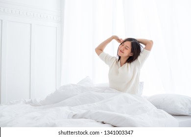 Asian Woman Wake Up In The Morning, Sitting On White Bed And Stretching, Feeling Happy And Fresh