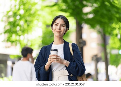 An Asian woman is walking on a green urban street with a relaxed expression, holding a cup of coffee. - Shutterstock ID 2312101059