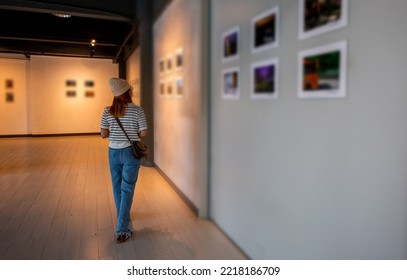 Asian woman walking through art gallery collection in front framed paintings pictures on white wall, Visitor person walk looking to picture at photo frame at interior show exhibition artwork gallery