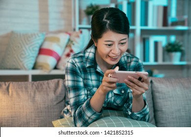 Asian Woman Watching Movie On Mobile Phone At Home In Night. Young Girl Having Fun Stay Up Late Enjoy Comedy Show Film Funny On Cellphone Sitting On Couch. Happy Female Laughing Using Technology.