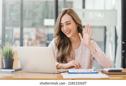 Asian Woman Waving Hello Talking On A Video Call. Successful Young Woman Sitting At Business Workspace. Business Conference Via Laptop.