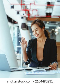 Asian Woman Waving Hello Talking On A Video Call. Successful Young Woman Sitting At Business Workspace. Business Conference Via Laptop.