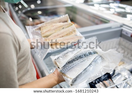 Asian woman wear face mask,choosing packed frozen cut fish in freezer in food department of supermarket,people panic buying, hoarding during the Covid-19,Coronavirus spread,girl preparing for pandemic