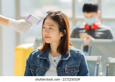 Asian Woman Wearing Blue Jean Shirt Sitting In The Airport Terminal Getting Temperature Check By Infrared Thermometer Or Temperature Gun. Coronavirus Covid-19 Concept.