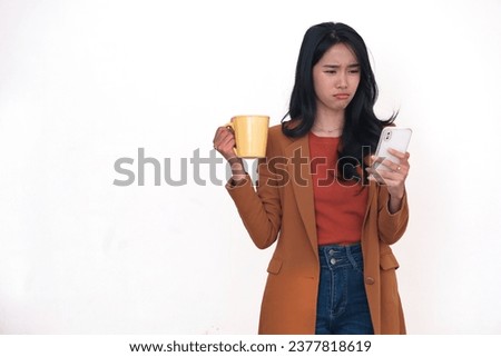 An Asian woman wearing a dark blazer is  checking her smartphone while holding a yellow cup; pouting, sad expression