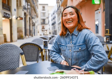 Asian woman wearing denim jacket laughs and enjoys her time at an outdoor cafe in valencia, spain - Powered by Shutterstock - Get 15% off with code: PIKWIZARD15