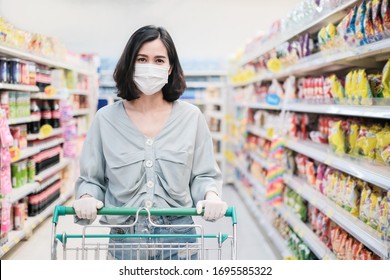 Asian Woman Wearing Face Mask And Rubber Glove Push Shopping Cart In Suppermarket Departmentstore. Girl Choosing, Looking Grocery Things To Buy At Shelf During Coronavirus Crisis Or Covid19 Outbreak.