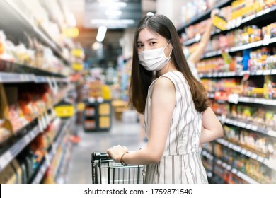 Asian Woman Wearing Face Mask Push Shopping Cart In Suppermarket . Girl Choosing, Looking Somethings To Buy At Shelf During Coronavirus Crisis Or Covid19 .happy Lifestyle Or Shopaholic Concept.