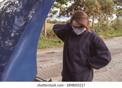 An Asian Woman Wearing A Face Mask Scratches The Side Of Her Head In Frustration As Her Car Broke Down. The Hood Of The Vehicle Is Raised Up.