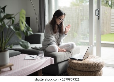 Asian Woman Wearing Glasses Laughing And Waving At Her Work Colleagues During A Video Call In Her Laptop Computer While Working From Home Sitting On A Sofa In The Living Room Of A House In Edinburgh