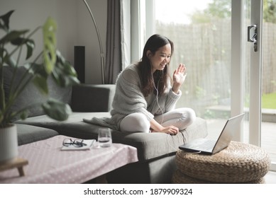 Asian Woman Wearing Glasses Smiling And Waving At Her Work Colleagues During A Video Call In Her Laptop Computer While Working From Home Sitting On A Sofa In The Living Room Of A House In Edinburgh,UK