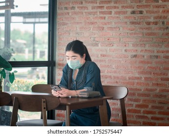 An Asian Woman Wearing A Green Mask Is Sitting, Using A Telephone On The Dining Table In A Restaurant And Having A Concrete Background.