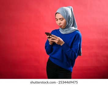 An Asian woman wearing a grey hijab and blue sweater is looking at her smartphone with a focused expression. She is standing against a red background, concentrating on the screen. - Powered by Shutterstock