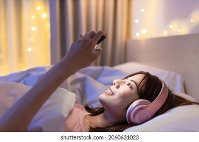 asian woman wearing headphone play mobile games by smartphone while lying on bed at night - Powered by Shutterstock