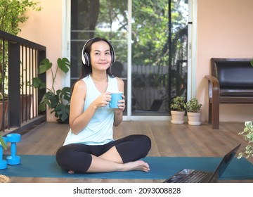 Asian Woman Wearing Headphones, Holding Coffee Cup, Sitting On Yoga Mat In Balcony  With Computer Laptop And Dumbbell, Smiling And Looking Up Copy Space, Thinking Creative Idea. Yoga Training Online